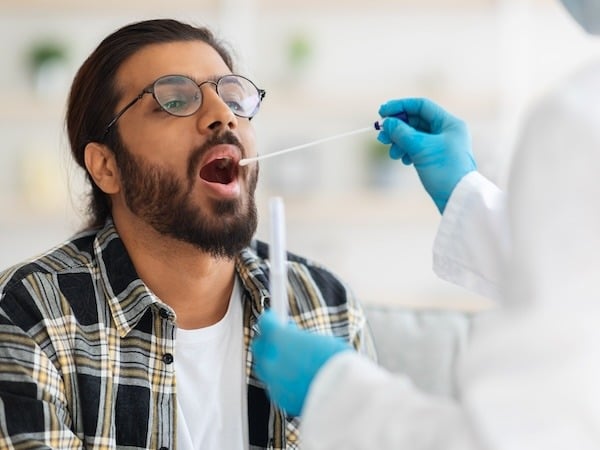 A healthcare professional wearing blue gloves and a white coat administers a swab test to a bearded man with glasses and a plaid shirt. The man is seated and has his mouth open while the swab is being taken.