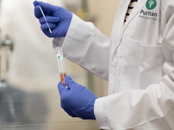 A lab technician wearing a white lab coat with a Puritan logo and blue gloves holds a sterile swab and a vial containing viral transport media in a laboratory setting.