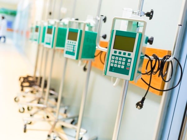  row of green and white infusion pumps mounted on IV stands in a hospital hallway. The pumps are aligned and connected with cables, emphasizing medical equipment used for delivering fluids or medications to patients.