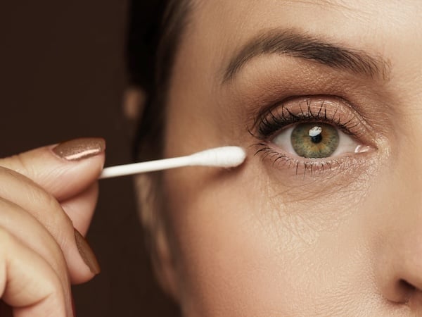 A woman holds a swab up to her eye to indicate makeup application. 