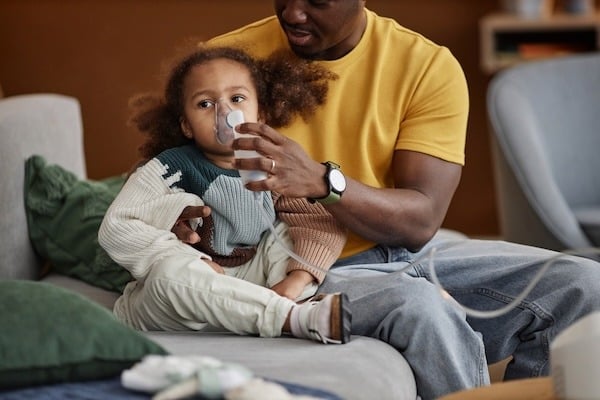 A young child sitting on a couch, holding a nebulizer mask to their face while an adult, seated next to them, gently supports the child. The adult is wearing a yellow shirt and jeans, while the child is dressed in a cozy, color-blocked sweater and pants. The scene suggests the child may be receiving respiratory treatment.