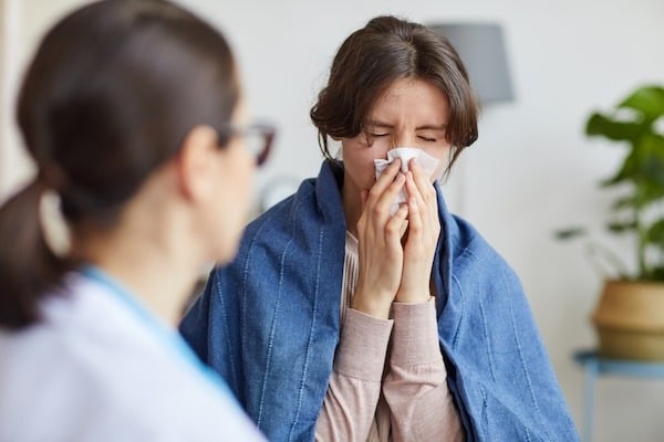 A woman sitting indoors, wrapped in a blue blanket, sneezes or blows her nose into a tissue while a healthcare professional, partially visible in the foreground, listens attentively. The scene suggests the woman may be experiencing cold or flu symptoms.