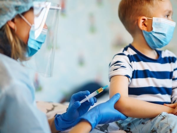 A healthcare worker wearing a face mask, face shield, and blue gloves administers a vaccine injection to a young child. The child, also wearing a face mask, looks away as they sit calmly in a striped shirt.