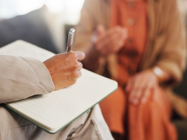 A close-up image of a person holding a pen and writing in a lined notebook while sitting across from another individual, who is gesturing with their hands during a conversation. Both are dressed in neutral-toned clothing, suggesting a professional or counseling setting.