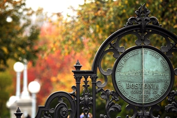 Ornate wrought iron gate featuring the official seal of the City of Boston, inscribed with the Latin phrase “Sicut Patribus Sit Deus Nobis” and the name “BOSTONIA.” The background shows autumn foliage in warm reds and oranges, with a soft focus on historic streetlamps.