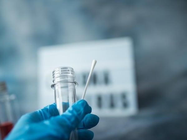 A gloved hand holds a sterile cotton swab and an open transparent sample collection tube. In the blurred background, additional sample vials with red liquid and a white sign with black text are visible, suggesting a laboratory or medical testing setting.