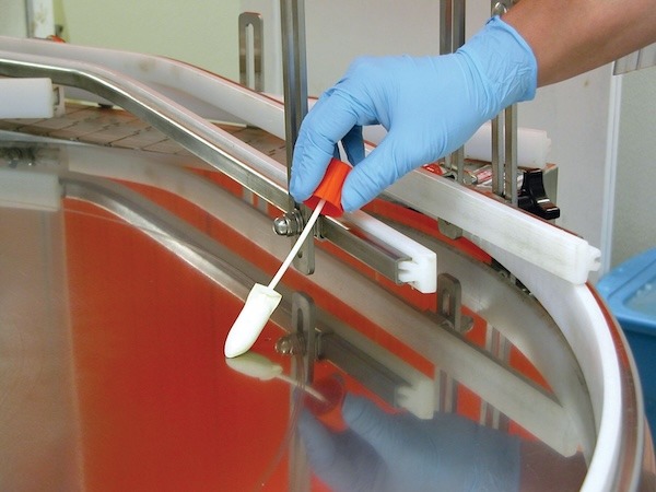 A gloved hand holds a swab while collecting a sample from the surface of a metal or plastic workspace area, likely for environmental testing or contamination checks.