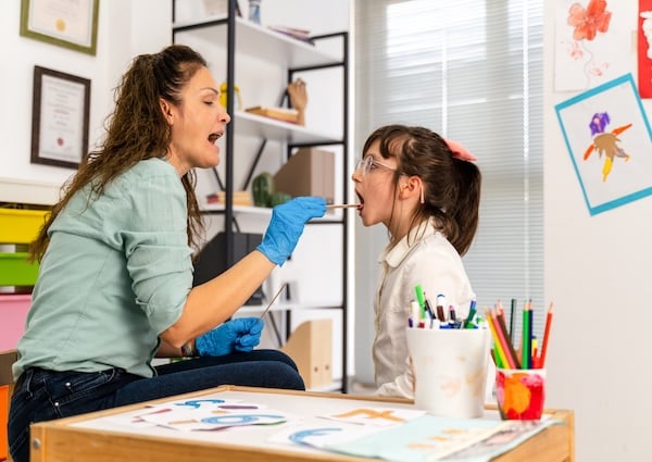 A speech therapist in a clinical setting, wearing blue gloves and a casual teal blouse, is conducting a speech articulation exercise with a young girl. 
