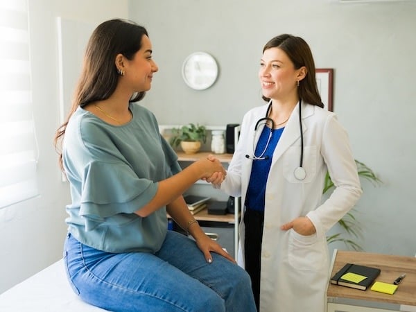 Female doctor smiling and shaking hands with a female patient who is sitting on an exam table in a medical office, suggesting a positive and supportive interaction.