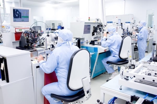 Technicians in full-body cleanroom suits operate precision equipment and microscopes in a high-tech laboratory environment. The workspace is sterile, with white walls, bright lighting, and specialized machines used for microelectronics or biomedical manufacturing.