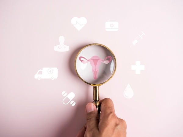 A hand holding a magnifying glass focuses on an illustration of a uterus against a soft pink background. Around the magnifying glass, there are white medical icons, including a heart, syringe, pills, a doctor silhouette, and a first aid cross, symbolizing women's health and gynecological care.